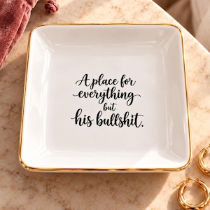 May include: A square white ceramic trinket dish with a gold-toned rim. The center features black cursive text that reads: A place for everything but his bullshit. The dish is accompanied by a dusty rose velvet pouch and two gold hoop earrings.