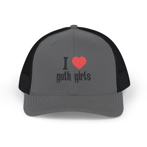 May include: A charcoal gray trucker hat with a black mesh back and a structured front panel. The front features an embroidered design with the text 'I' in black, a red heart symbol, and the text 'hot girls' in a stylized black font.