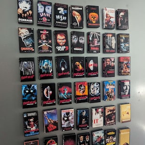 Könnte beinhalten: Eine Sammlung von zweiundvierzig rechteckigen VHS-Videokassetten, die in einem Raster an einer Wand angeordnet sind. Die Kassetten zeigen verschiedene Horror- und Actionfilm-Cover, darunter Titel wie Scream, Halloween, Child's Play und Terminator.