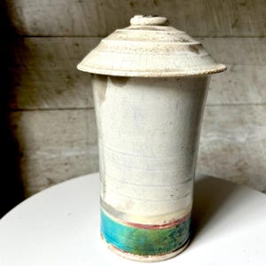 May include: A tall, off-white ceramic vase with a matching lid. The vase has a cylindrical shape with a turquoise, red, and green band near the base. The lid is tiered and slightly weathered, giving it a rustic appearance. The vase is sitting on a white surface.