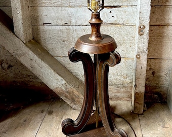 May include: A vintage wooden table lamp with a bulb. The lamp has a dark brown, ornate base with curved supports and a round top. The lamp's metal arm holds a light bulb. The lamp is on a wooden floor.