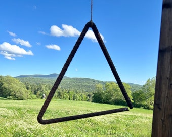 Puede incluir: Un triángulo de metal colgando frente a un campo verde con un cielo azul y nubes blancas en el fondo.