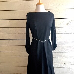 May include: A black dress with a boat neck and long sleeves. The sleeves have a lace pattern. The dress has a silver chain belt.