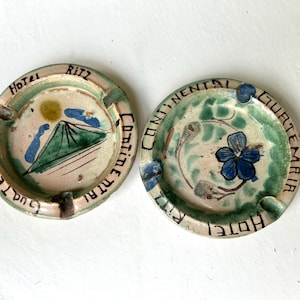 Puede incluir: Dos ceniceros de cerámica vintage con diseños pintados a mano. Uno presenta una escena de montaña y las palabras "Hotel Ritz Guatemala Continental". El otro tiene una flor azul y el mismo texto. Ambos tienen un esquema de color verde y crema.