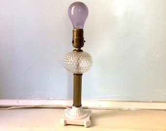 May include: A vintage table lamp with a white ceramic base and a clear glass shade with a textured surface. The lamp has a brass stem and a purple light bulb.