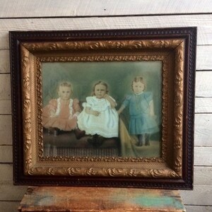 May include: An antique picture frame with a gold-colored ornate border and a dark brown wood exterior. The frame contains a vintage photograph of three young children in dresses, sitting on a bench.