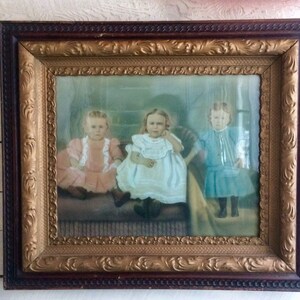 May include: An antique framed photograph of three young girls in white, pink, and blue dresses. The girls are sitting on a couch and smiling at the camera. The frame is ornate with gold accents.