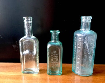 May include: Three antique glass bottles with a light green tint. The bottles are rectangular in shape and have a rounded top. The middle bottle has the text "INDIAN ROOT BEER EXTRACT" embossed on the front. The bottle on the right has the text "VANDICE'S MUSTARD  PREPARED  AT WOOD  CINCINNATI, OHIO" embossed on the front.