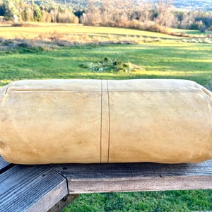 May include: A large, cylindrical, tan-colored leather bag with a stitched seam down the center. The bag is resting on a weathered wooden surface, with a green field and trees in the background.