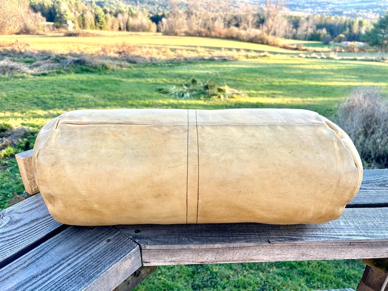 May include: A large, cylindrical, tan-colored leather bag with a stitched seam down the center. The bag is resting on a weathered wooden surface, with a green field and trees in the background.