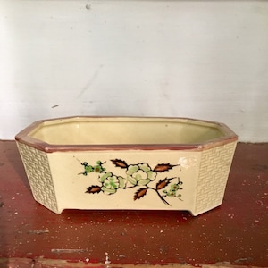 May include: Cream-colored rectangular planter with a floral design. The planter has a woven texture on the sides and a brown rim. The floral design features green and white flowers with brown stems and leaves.