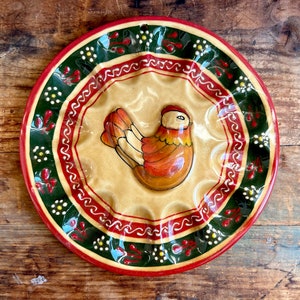 Könnte beinhalten: Ein Keramik-Eierhalter in Form eines Tellers mit rotem, grünem und weißem Rand. Die Mitte des Tellers zeigt ein braunes und oranges Hühnermotiv.