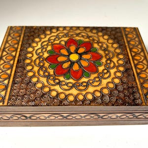 May include: Rectangular wooden box with a floral design. The lid features a central flower in red, orange, and yellow, surrounded by intricate scrollwork. The box has a light brown base with decorative borders, showcasing a blend of natural wood tones.