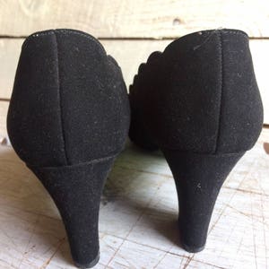 May include: A pair of black suede heeled shoes with a scalloped edge detail. The shoes have a pointed toe and a high heel.