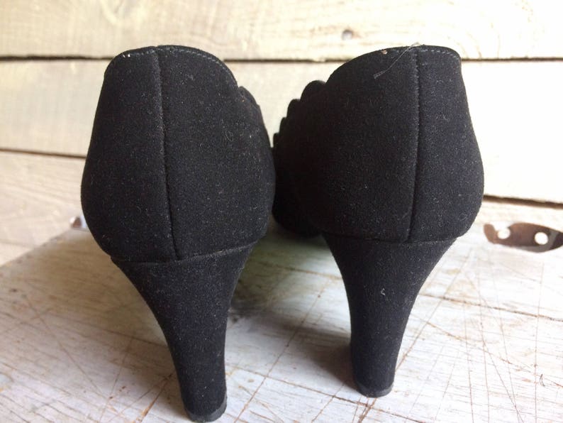 May include: A pair of black suede heeled shoes with a scalloped edge detail. The shoes have a pointed toe and a high heel.