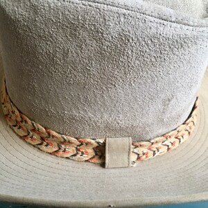 May include: A gray suede cowboy hat with a braided band featuring red, black, and white accents.