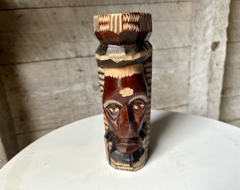May include: A wooden carving of a person's face with intricate details. The carving is brown and white and has a geometric pattern. The face is carved with a serious expression.