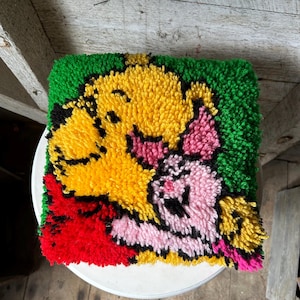 May include: A colorful, hand-stitched rug featuring a yellow dog and a pink pig hugging. The rug is made of yarn and has a green background.