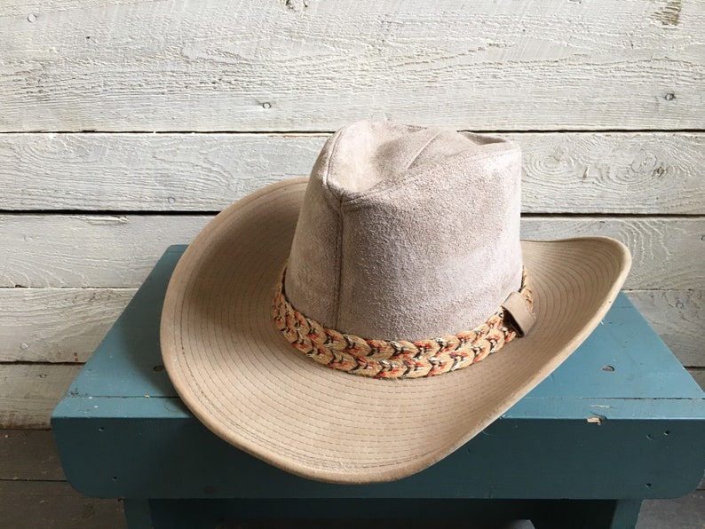 May include: A light brown suede cowboy hat with a braided band featuring orange and brown accents. The hat is sitting on a blue surface.