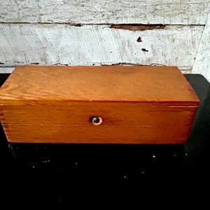 May include: A rectangular wooden box with a warm, medium-brown finish. The box has a small, round metal knob centered on the front. The box is sitting on a black surface, with a weathered white wooden background.