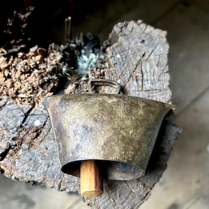 May include: A weathered brass cowbell with a wooden clapper, resting on a rough-hewn wooden surface.