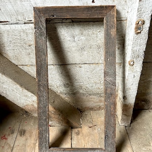 Op de afbeelding: Een rechthoekige fotolijst gemaakt van verweerd, hergebruikt hout met een natuurlijke grijsbruine afwerking. De lijst heeft een zichtbare houtnerf, knoesten en een ruw, getextureerd oppervlak met in verstek gezaagde hoeken.