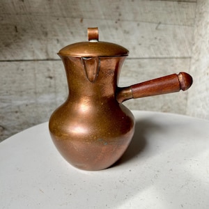 May include: A copper coffee pot with a wooden handle and lid. The pot has a bulbous body and a spout for pouring. The handle is attached to the side of the pot. The lid has a small handle on top. The pot is made of copper and has a shiny finish.