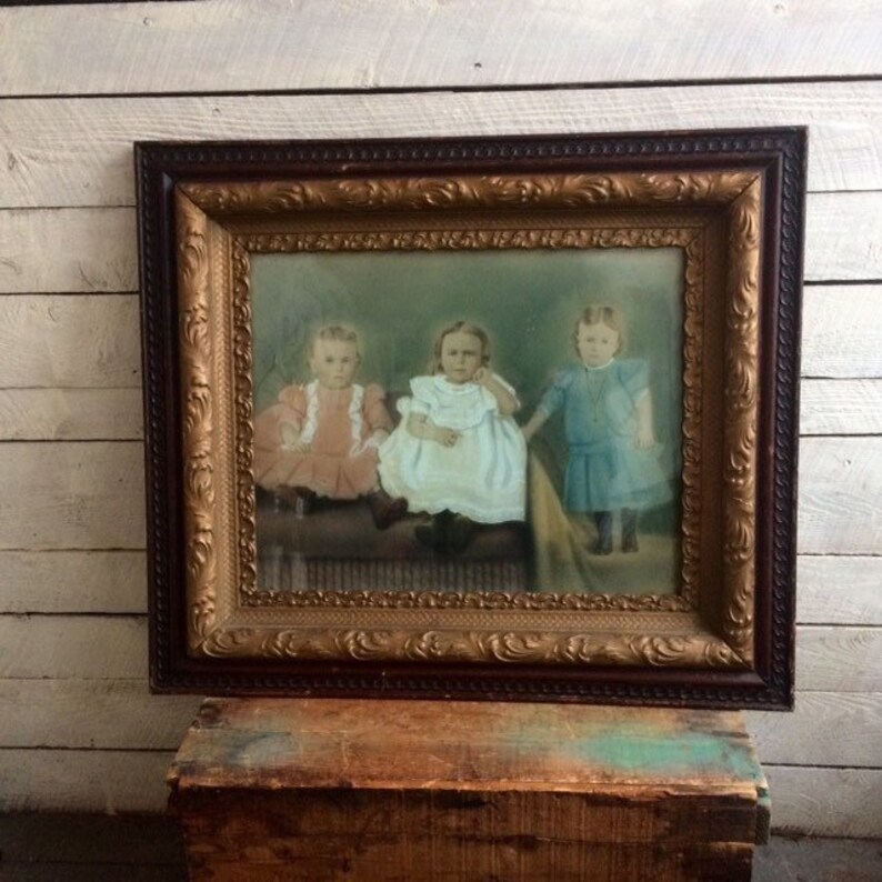 May include: An antique picture frame with a gold-colored ornate border and a dark brown wood exterior. The frame contains a vintage photograph of three young children in dresses, sitting on a bench.