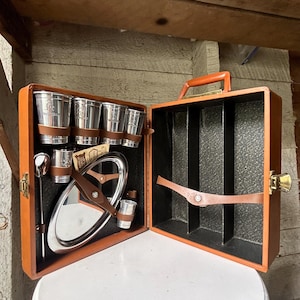 May include: A portable bar set in a tan rectangular case with a top handle and gold-tone latches. The interior features black textured lining, brown leather straps, four silver-tone metal cups, a silver-tone oval tray, and a long metal spoon.
