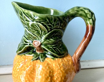 May include: A ceramic pitcher shaped like a lemon with green leaves and a brown handle. The pitcher is glazed in yellow, green, and brown.