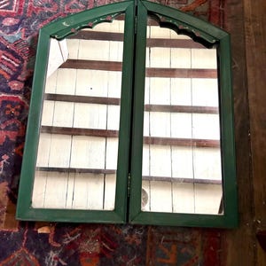 Puede incluir: Un mueble verde con la parte superior arqueada y dos puertas con espejo. El mueble tiene un adorno decorativo con detalles en rojo y blanco. El interior presenta estantes de madera blancos. El mueble está sobre una alfombra estampada y una pared de madera.