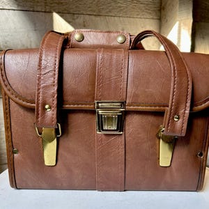 May include: A brown vintage briefcase with a rectangular shape. The briefcase has a handle, a clasp, and gold-colored metal accents. The exterior is made of a textured material, and the overall design is classic and functional.