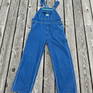 May include: Blue denim overalls featuring a front bib pocket with a green and yellow logo patch, adjustable shoulder straps with metal buckles, side button closures, and wide pant legs with visible stitching along the seams and leg panels.