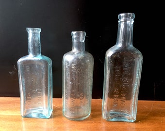 May include: Three vintage glass bottles with embossed lettering. The bottles are clear and have a light blue tint. The bottles are rectangular in shape and have a rounded bottom. The bottles are sitting on a wooden surface.