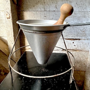 Puede incluir: Un colador de alimentos cónico de metal plateado con superficie perforada, que cuenta con un mango largo y un soporte de metal a juego. Un mortero de madera de color claro y superficie lisa se coloca dentro del colador para presionar alimentos.