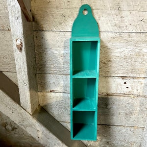 May include: A teal green wooden wall shelf with three shelves. The shelf has a decorative top with a hole in the centre.