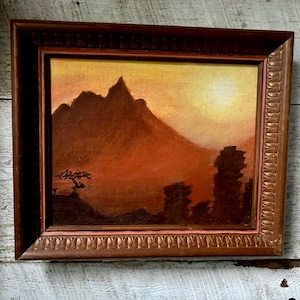 May include: An original landscape painting in a brown wooden frame. The artwork depicts a mountain range silhouetted against a sunset sky. The color palette is warm, with shades of orange, brown, and yellow. A small tree is visible in the foreground.