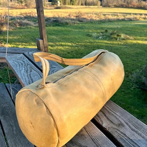May include: A tan cylindrical duffel bag with a zipper closure and a striped handle. The bag is made of a worn, textured material and is resting on a weathered wooden surface. The background features a green field and trees.