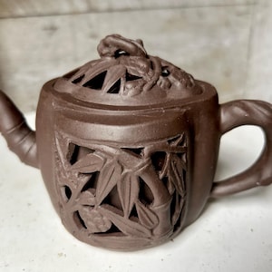 Op de afbeelding: Een bruine theepot van keramiek met een deksel met een gesneden bamboe ontwerp. De theepot heeft een tuit en een handvat.