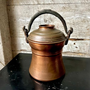May include: An antique copper-coloured pot with a lid and a black handle. The pot has a rounded body and a cylindrical neck. The handle is arched and attached with metal loops. The pot is on a black surface.