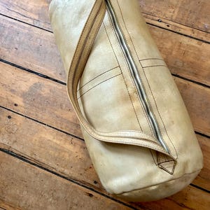 May include: A cylindrical, tan leather duffel bag with a full-length zipper and a single shoulder strap. The bag has visible stitching and a worn appearance, suggesting vintage use. The bag is resting on a wooden surface.