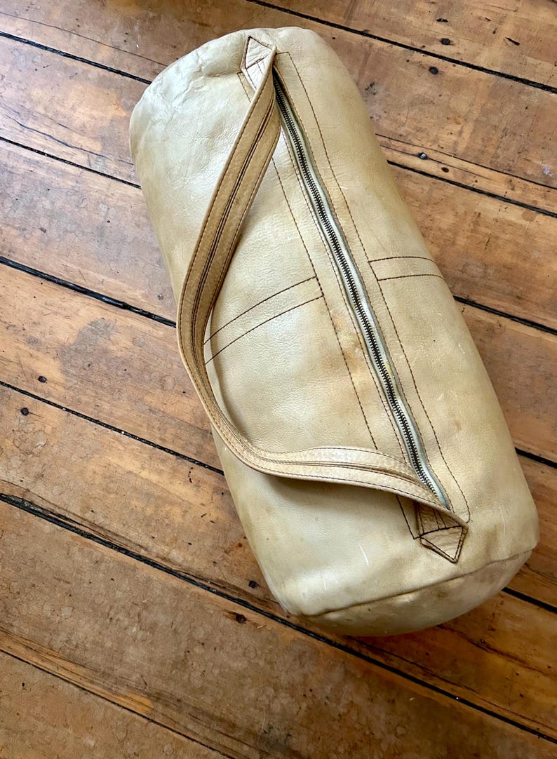 May include: A cylindrical, tan leather duffel bag with a full-length zipper and a single shoulder strap. The bag has visible stitching and a worn appearance, suggesting vintage use. The bag is resting on a wooden surface.
