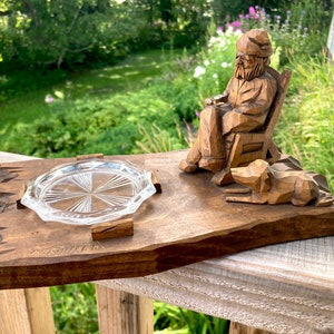 May include: A wooden sculpture of a man sitting in a chair with a dog at his feet. The sculpture is on a wooden base with a glass coaster in the centre. The sculpture is carved in a rustic style and has a natural wood finish.