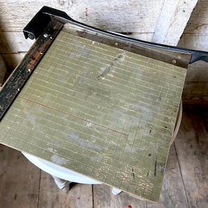 May include: A vintage paper cutter with a black handle and a green cutting mat with a grid pattern. The mat has a wooden ruler with the word "MONARCH" printed on it.
