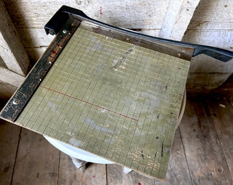 May include: A vintage paper cutter with a black handle and a green cutting mat with a grid pattern. The mat has a wooden ruler with the word "MONARCH" printed on it.