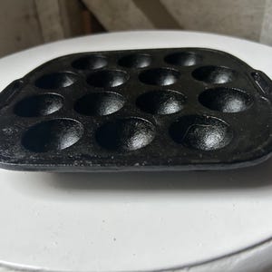 May include: Black cast iron takoyaki pan with 14 hemispherical molds. The pan has two small handles and is designed for cooking small, round Japanese octopus balls. The pan is on a white surface.