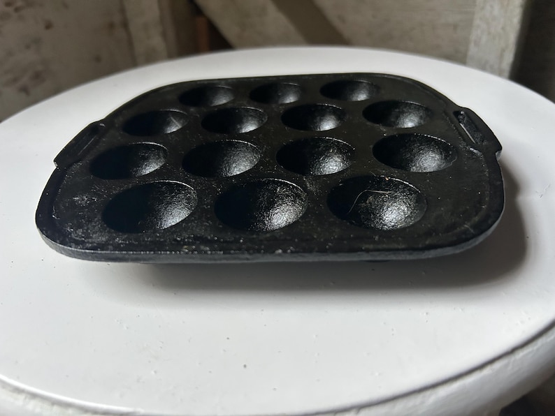 May include: Black cast iron takoyaki pan with 14 hemispherical molds. The pan has two small handles and is designed for cooking small, round Japanese octopus balls. The pan is on a white surface.