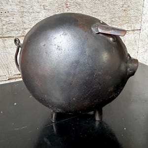 Puede incluir: Una hucha de hierro fundido oscuro, de forma redonda, con patas pequeñas y una cola enroscada. Tiene una ranura en la parte superior y un pequeño hocico. La superficie tiene una textura ligeramente rugosa y un acabado mate oscuro.