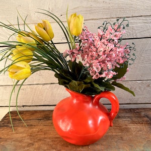 May include: A bright orange ceramic pitcher with a wide mouth and a handle, filled with a bouquet of yellow tulips and pink flowers.