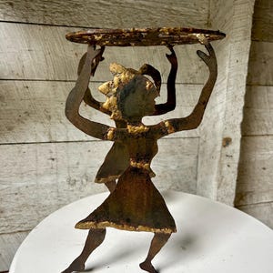 May include: A rustic metal sculpture of two figures holding up a circular tray. The figures are silhouetted in a dark brown color, with a textured, aged appearance. The tray is also brown and has a rough, uneven edge. The sculpture is placed on a white surface.